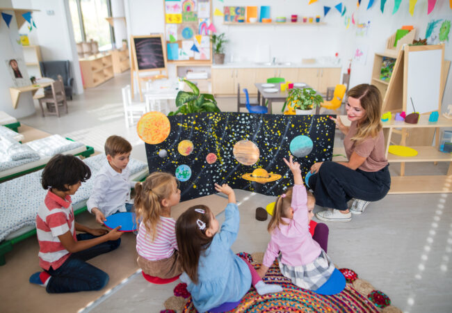 Multiracial group of small children with teacher sitting on floor and learning about space together at kindergarten.